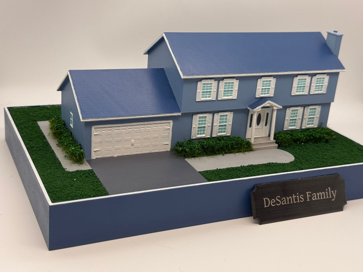 Photograph of a completed hand-painted blue-gray Colonial house miniature with white shutters, garage, landscaping, a plain printed base and personalized family plaque.