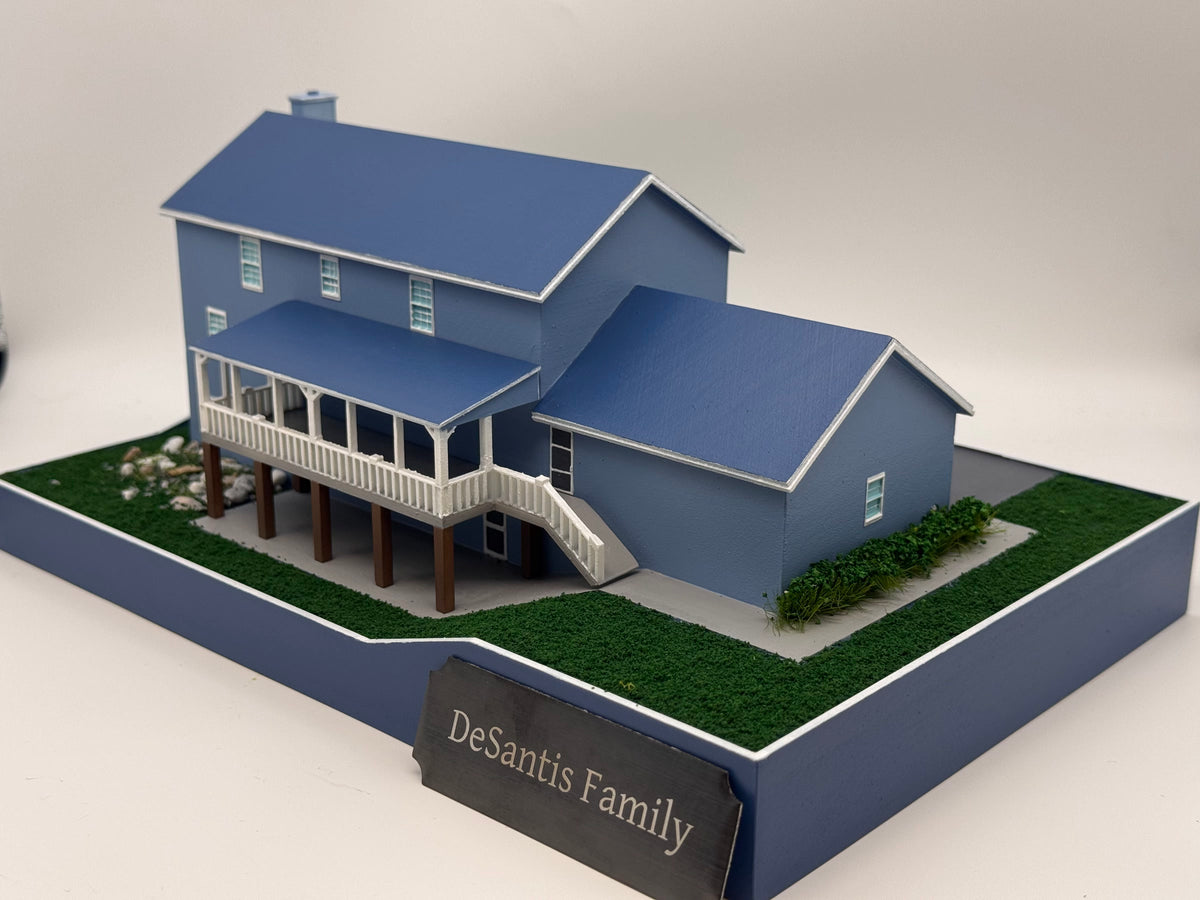 Photograph of the same completed blue-gray house miniature from the rear, with an elevated covered sunroom, white railings, supports, stairs, landscaping and plain printed base.