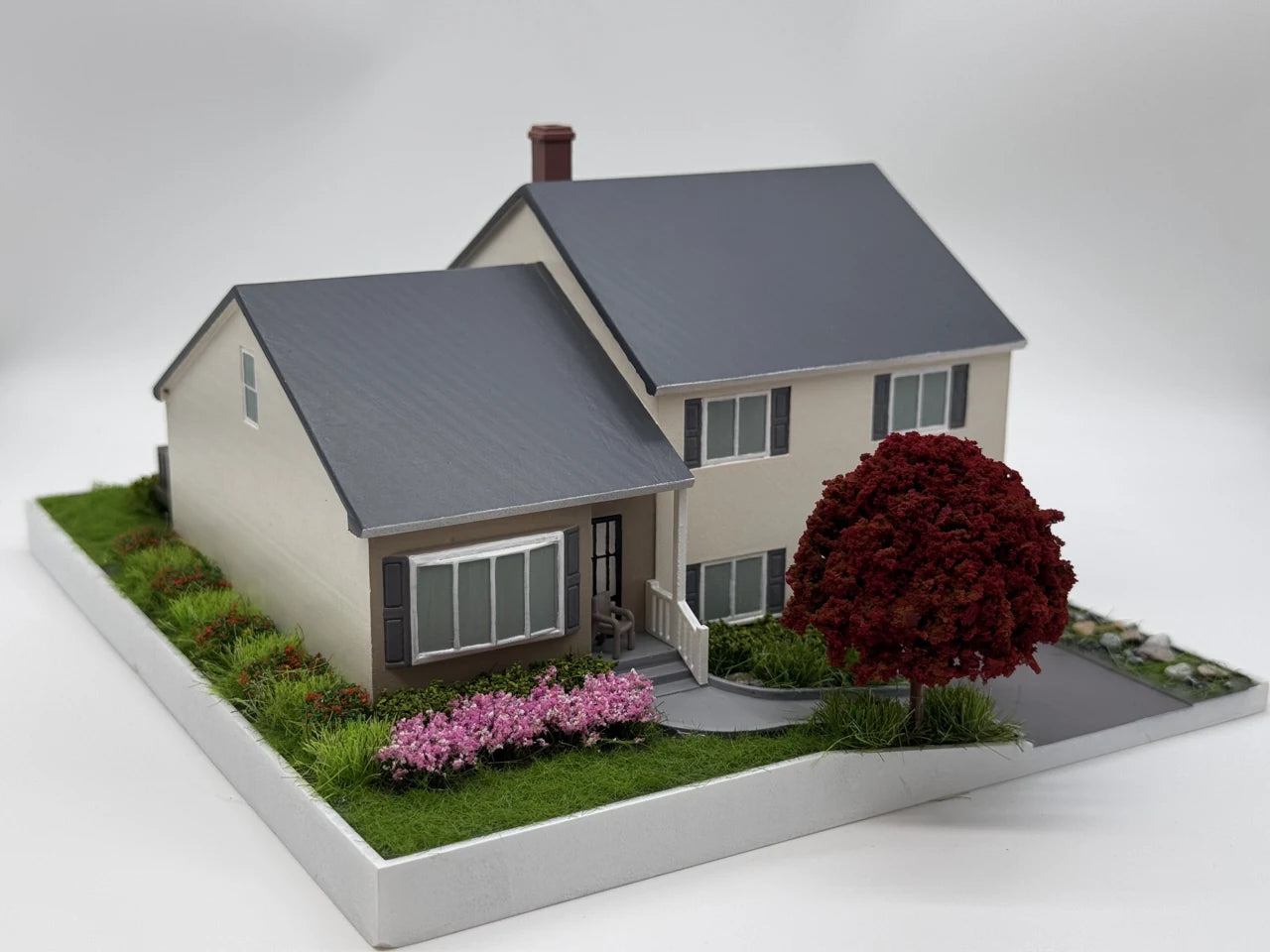 Photograph of the same family home as a completed model with cream walls, grey roofs, red-leaf tree and flowers.