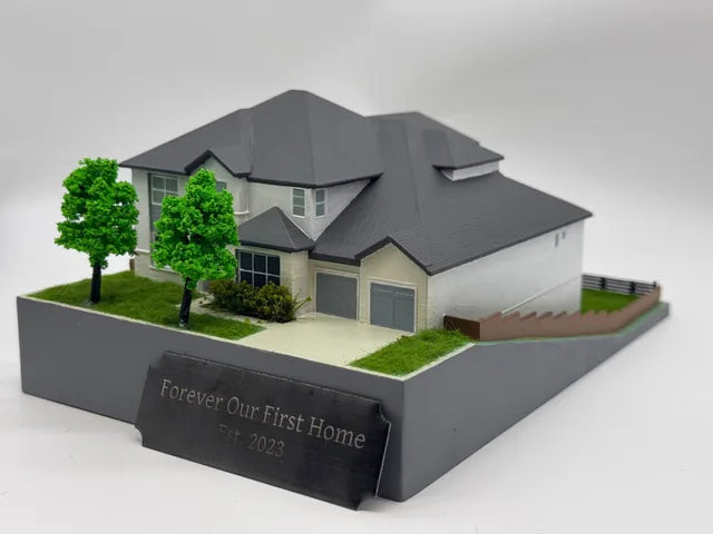 Completed model of a white two-storey home with grey hip roofs, a two-car garage, two trees, side fencing and a plaque reading Forever Our First Home, Est. 2023.